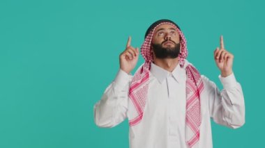 Orta Doğulu bir adam yukarıdan yukarıya bakıyor, stüdyoda yukarı doğru işaret ediyor. Geleneksel Arap kıyafetleri giyen genç İslamcı, başörtüsü ve cübbe giyiyor..