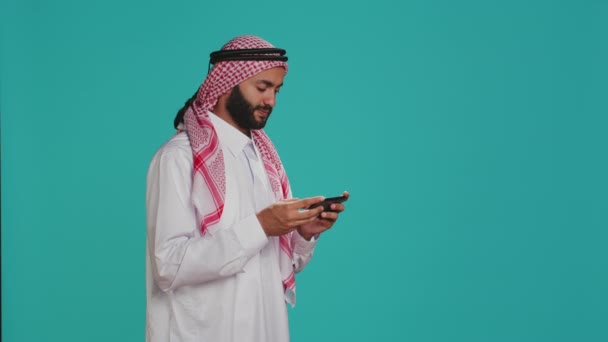 穆斯林男子穿着传统服装 在智能手机上玩电子游戏 站在蓝色背景之上 阿拉伯人在电话应用 娱乐等方面享受着手机游戏竞赛 — 图库视频影像