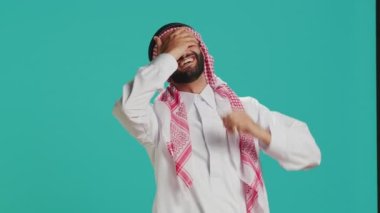 Stüdyoda şakaya gülen neşeli insan, geleneksel Müslüman kıyafetleri ve kufiye giyerken eğleniyor. Genç Arap adam insanlarla gülmekten zevk alıyor, mutluluk ve neşe ifade ediyor..