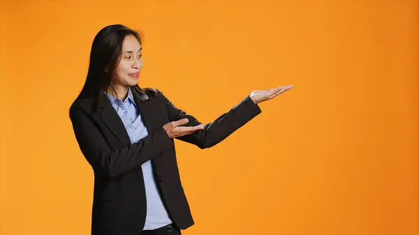 Asian employee points at left or right sides in studio, creating new web commercial over orange background. Young business manager working on marketing advertisement during shoot.