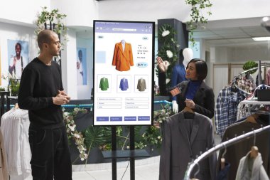Asyalı mağaza asistanı, giyim mağazasından moda koleksiyonu giysileri seçerek, self servis büfesinin önündeki adama eşyalar gösteriyor. Beyaz müşteriler kadın çalışanlarla ürünleri tartışıyor.