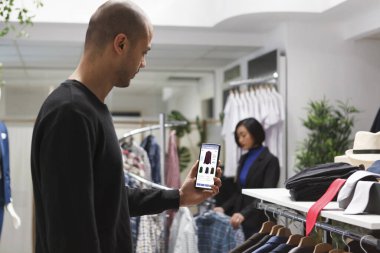 Kafkasyalı bir adam butik mağazasının web sitesini gösteren mobil bir cihaz tutuyor. Giysi raflarının yanında duran genç erkek müşteri, tanıtım amaçlı kıyafet fiyatlarını internette gösteren bir akıllı telefonu kavrıyor..