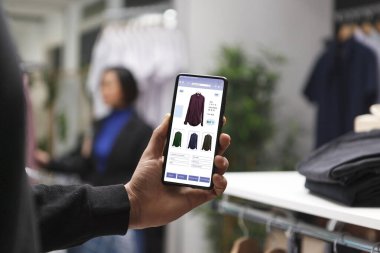 Bir giyim mağazasındaki beyaz bir müşteri elinde akıllı bir telefon tutuyor ve bir butik web sitesinden kıyafetlere bakıyor. Erkek alışverişi, alışveriş merkezindeki giysi parçalarını cep telefonuyla incelemektir..