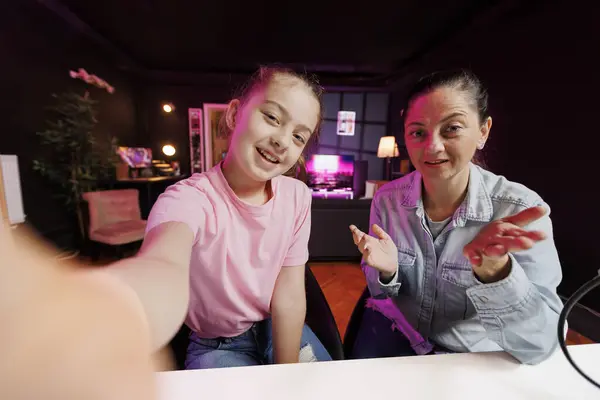 Lively child and her mum do content creation using selfie camera to film vlog channel intro. Daughter and parent greet fanbase, presenting today video topic in pink neon illuminated studio, POV shot