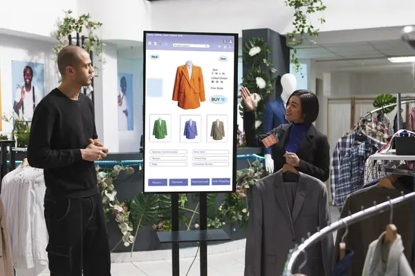 stock image Asian store assistant showcasing items to man in front of self service kiosk, selecting fashion collection clothes from clothing boutique. Caucasian customer discussing products with female employee.