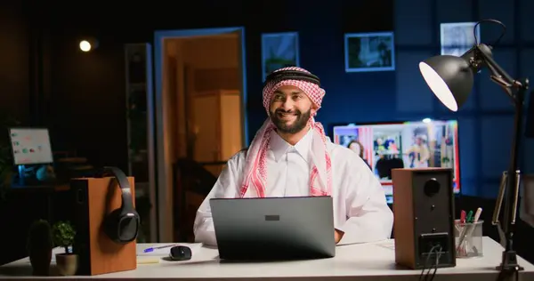 笑顔のアラブのテレワーカーはスタイリッシュなアパートで仕事の電子メールに答え すぐに仕事を終えることに満足しています 穏やかなイスラム教徒の従業員はリモートで働き バックグラウンドノイズとして開かれたテレビでラップトップ上のデータを入力します — ストック写真