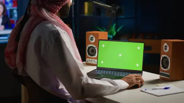 アラビア人男性が自宅で仕事をしている間 モックアップラップトップをスクロール バックグラウンドノイズとしてテレビ付きのアパートオフィスの隔離された画面デバイスに電子メールを書く自己雇用者 — ストック写真