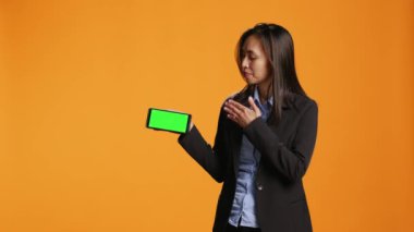 İş kadını elinde yeşil ekran şablonu olan bir telefon tutuyor, turuncu arka planda poz veriyor ve stüdyoda boş bir telif alanı gösteriyor. Akıllı telefon uygulamasında krom ekranlı Asyalı yetişkin.