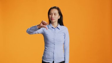 Asyalı bir kadın kameraya karşı tavır sergiliyor. Turuncu arka planda anlaşmazlık ve olumsuzluk gösteriyor. Negatif mutsuz yetişkin, stüdyoda baş parmağını aşağı gösteren sembol, yanlış fikir..