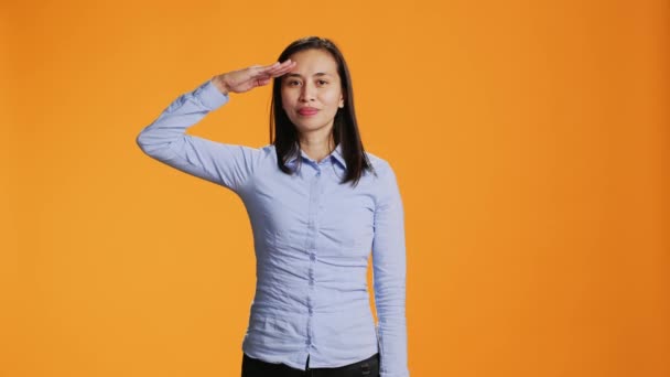 カメラに軍事的な敬礼のジェスチャーを示す愛国的な女性は 寺院の上に手を挙げ スタジオで誇りに思っています アジア人 オレンジ色の背景に対する軍のシンボルで塩漬け — ストック動画