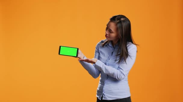 自信的女孩在手机显示屏上显示绿色 站在橙色的背景上 在设备上显示空白的铬色键 亚洲人手持带有独立模型的智能手机屏幕 — 图库视频影像