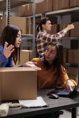 Depo işletmecisi, iş arkadaşının depoda dizüstü bilgisayarda envanter çıkarmasını denetliyor. Depoda genç Asyalı kadın çalışanlar mal lojistik ve dağıtım programını tartışıyor