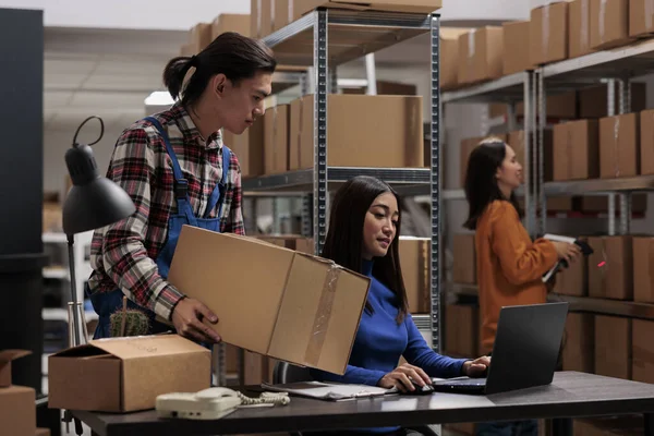Warehouse asian employees managing ecommerce order fulfillment in storage room. Storehouse carrier holding cardboard box with goods while logistics manager planning delivery on laptop