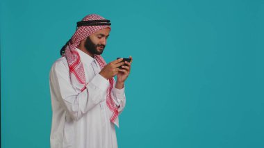 İslami adam telefonda eğlenceli mobil video oyunları oynuyor. Geleneksel başörtüsü takmış ve türban takıyor. Ortadoğulu kişi akıllı telefon uygulaması kullanarak online oyun yarışmasına katılıyor..