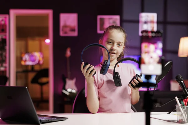 Kid presenting headphones in different price ranges to audience, comparing them to find winner, filming with phone on tripod. Young tech content creator recommending listening devices to fans