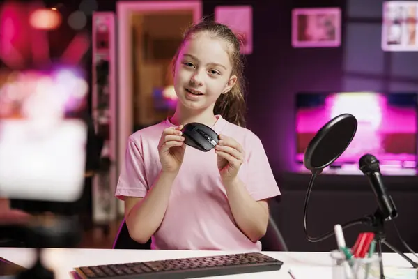 stock image Cute girl in studio reviewing gaming mouse in front of camera in neon lit studio. Joyous youngster filming electronics haul, presenting wireless computer peripheral to viewers