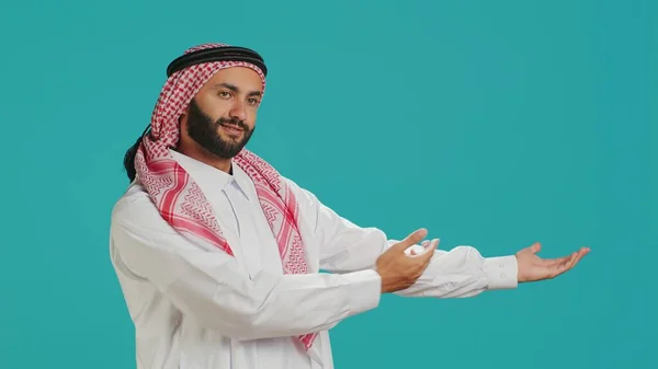 Muslim guy wearing traditional robe and ghutra gesturing to side with hands and posing for studio shot. Arabic smiling person promoting product while showing marketing ad aside.