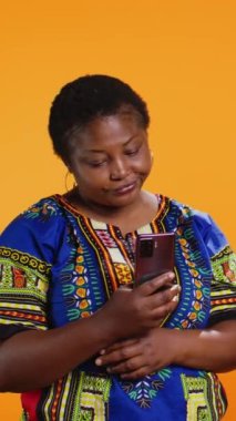 Geleneksel giysiler içinde dikey Video Etnik insanlar akıllı telefon uygulamalarını kontrol ediyor, sosyal medya ağından gelen mesajlara bakıyor. Afrika kökenli Amerikalı erkek ve kadın mobil cihazda web sitelerine bakıyorlar.