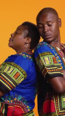 Dikey Video Ciddi gururlu etnik çift arka arkaya stüdyoda duruyor, turuncu arka planda kendilerine güveniyorlar. Afrikalı erkek ve kadın geleneksel giyinmiş, tasasız.