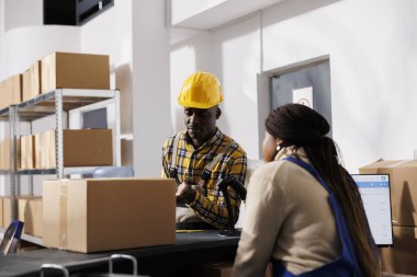Afrika kökenli Amerikalı erkek ve kadın depo çalışanları depo masasında sohbet ediyorlar. Posta servisi çalışanları posta masasında paket sevkiyatını tartışıyor.