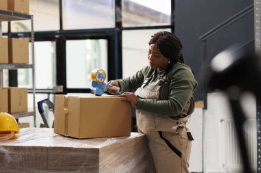 Afro-amerikan yönetici müşterileri sipariş hazırlıyor, ambardaki karton kutulardaki ürünleri yapıştırmak için yapışkan bant kullanıyor. Depo çalışanı depo envanteri sırasında endüstriyel kıyafetler giyiyor.