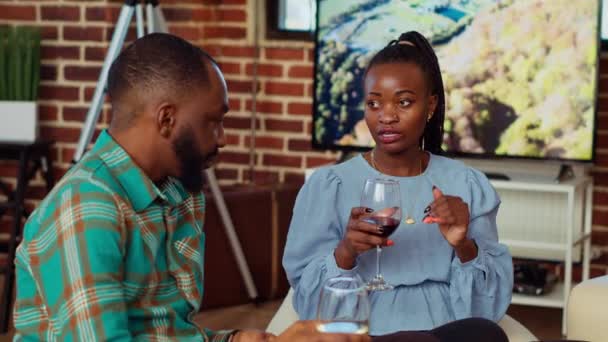 アフリカ系アメリカ人男性がソーシャルメディアでワインを飲みながらソファーでガールフレンドと話す ブリックウォールの家で楽しい会話を持つアパートパーティーで夫と妻ゲスト — ストック動画