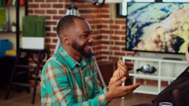 笑顔のアフリカ系アメリカ人男性は スナックを食べながらBipocアパートパーティーのゲストと面白い会話をしています 友人の会話の包括的なグループの隣のテーブル上のチャーターボードに閉じる — ストック動画