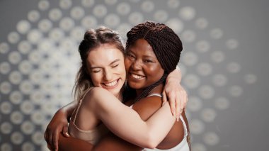 Cheerful gentle girls embracing imperfections on camera, posing together with self confidence. Interracial women with glowing skin feeling beautiful and unique in studio, advertising self love.