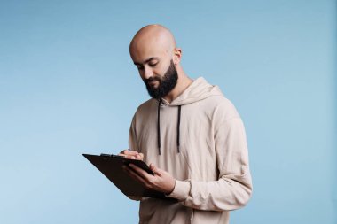 Konsantre Arap adam pano tutarken kontrol listesi hazırlıyor ve yazıyor. Gündelik kapüşonlu giyen, listedeki eşyaları kontrol eden ve stüdyoda notlar alan genç bir adam.