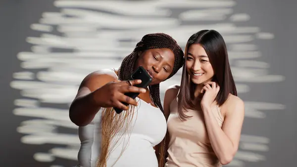 Two skincare models taking photos with smartphone, diverse young women advertising different skintones and body shapes. Beautiful luminous girls having fun with pictures for beauty ad.