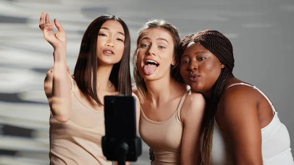 Interracial group of friends taking photos on smartphone to promote self acceptance and body positivity. Women laughing and acting funny on pictures, enjoying skincare ad campaign.