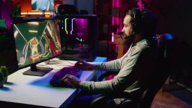 Mutlu adam karanlık oturma odasındaki çevrimiçi arkadaşlarıyla iletişim kurmak için kulaklık kullanıyor, bilgisayar masasında bilgisayar oyunları oynuyor, işten sonra dinleniyor.