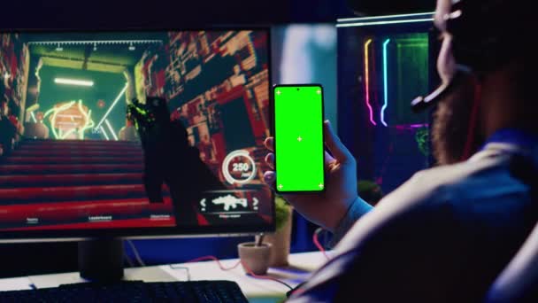 グリーンスクリーン携帯電話でチュートリアルを見ている男は 銃撃レーザー弾で最初の人を撃つビデオゲームをプレイしながら スマートフォンでオンラインガイドを見ることでゲームを学ぶGamer — ストック動画