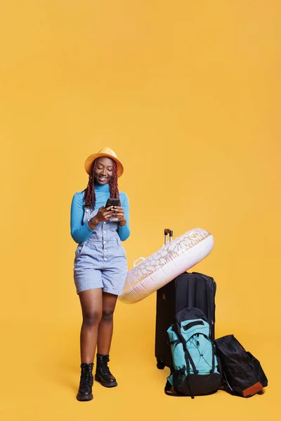 Young tourist checking smartphone app, posing with trolley bags and inflatable in studio. Female model feeling happy on inernational vacation, using mobile phone online browser.