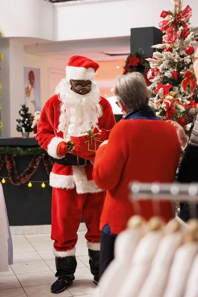 Santa Claus worker in suit inviting customer to participate in raffle contest to win free clothing items during christmas holiday festivity. Jolly decorations and xmas tree in store.
