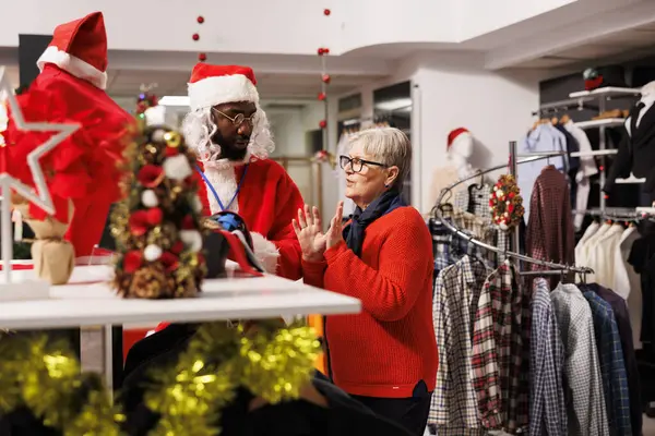 Employee in suit presenting clothes to woman, searching for fashion items as presents. Man portraying santa claus greeting customers and giving assistance during holiday sales.
