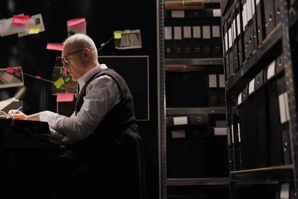 Elderly investigator working overtime at crime scene report, analyzing evidence files in arhive room. Overworked private detective checking criminology documents, planning strategy to catch suspect