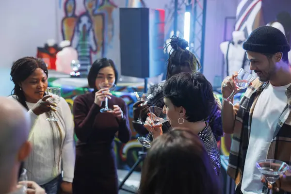 Diverse people crowd drinking alcohol beverages while celebrating in nightclub. Youn friends group holding glasses while socializing and partying on dancefloor at social gathering