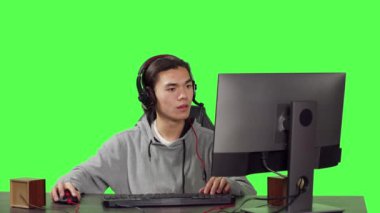 Genç oyuncu bilgisayar masasında oturuyor ve arkadaşlarıyla çevrimiçi video oyunları oynuyor, iyi vakit geçirmek için modern cihazlar kullanıyor. Asyalı adam yeşil ekran üzerinden roket turnuvasına katılıyor.