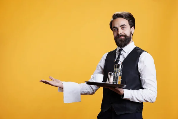 Professional server presenting something in studio, providing luxurious service at restaurant while he carries platter with cutlery and coffee. Male butler serving drinks to clients, fine dining.