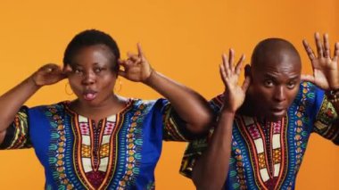 Komik suratlı aptal etnik çift stüdyoda eğleniyor ve dillerini dışarı çıkarıyorlar. Neşeli insanlar eğlenceli jestler yapıyor ve sette eğleniyorlar, turuncu arka plan.