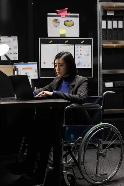 Paralyzed businesswoman accountancy executive in wheelchair adding budget plan data on laptop. Asian woman with paraplegia in storage office filled with paperwork folders on shelves and flowcharts