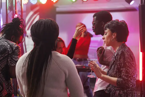 Young friends drinking alcohol and partying on crowded club dancefloor illuminated with spotlights. Diverse people dancing and enjoying nightlife activity at nightclub discotheque