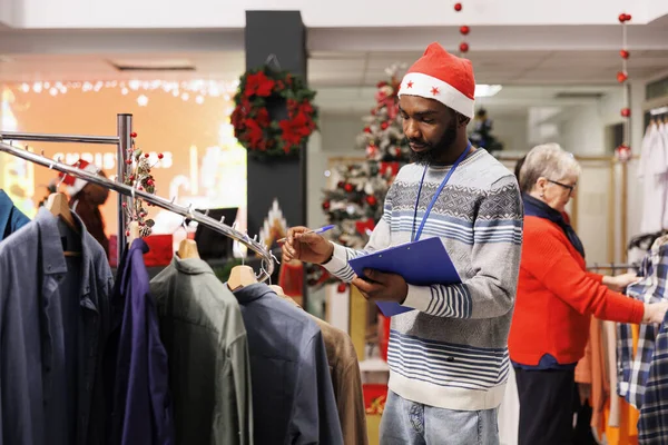Retail worker doing items inventory with clipboard in hand, wearing santa hat in clothing store decorated for christmas. Shopping assistant counting all merchandise and clothes from hangers.