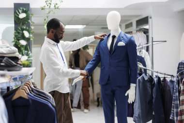 Butik asistanı, erkek giyim kuşamında mankenleri kontrol ediyor. Afro-Amerikalı bir adam alışveriş merkezinde çalışırken erkek lüks takım elbisesinin modelini inceliyor.