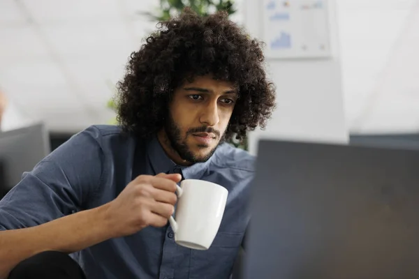 Entrepreneur drinking coffee while working on laptop to meet project deadline in business office. Arab start up company employee holding tea mug and researching data for analysis