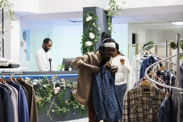 Afro-Amerikalı neşeli adam kulaklıkla müzik dinlerken kıyafet alışverişi yapıyor. Kulaklıklı mağaza müşterisi butikte kıyafet seçerken askıda tişört tutuyor.