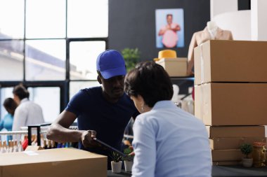 Afro-Amerikan kurye lojistik raporunu butikte müşteri nakliyesi detaylarını tartışırken işçiye gösteriyor. Çalışan teslimat için paketler hazırlıyor, alışveriş merkezinde online siparişlerle çalışıyor.