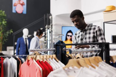 Afro-amerikan tarzı bir adam gündelik gömlekleri kontrol ediyor, modern butikte moda kıyafetler alıyor. Moda ürünleri kumaş ve giyim mağazasında fiyat analizi yapan alışveriş hastası
