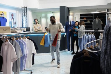 Neşeli Afrikalı Amerikalı adam alışveriş torbaları taşıyor ve giysi dükkanından alışveriş yaptıktan sonra dans ediyor. Moda butiği mutlu müşteri içi kıyafet dolu kağıt paketler tutuyor.